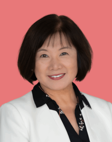 A woman with short dark hair wearing a white blazer over a black collared shirt, smiling at the camera, with a solid pink background.